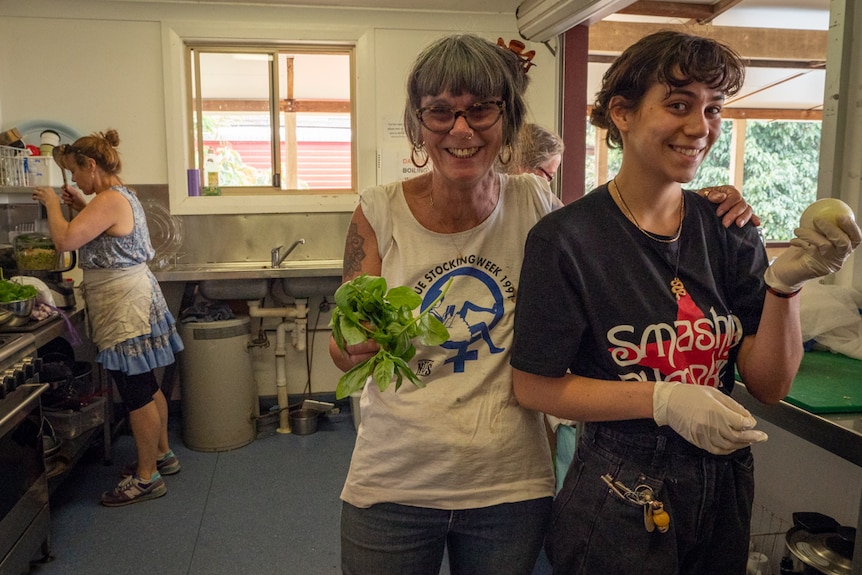 Two woman smiling at camera, standing in a kitchen in a hall, one holding basil and the other an onion.