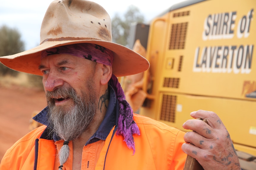 A man with a wide brimmed hat and a spider tattoo on his neck standing in front of a truck. 