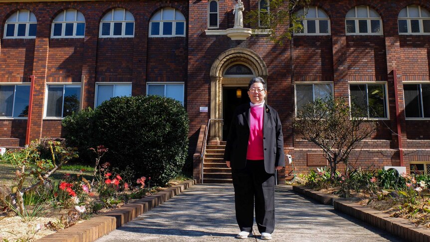 Josephite Sister Margaret Ng stands in front of brick religious building and garden.