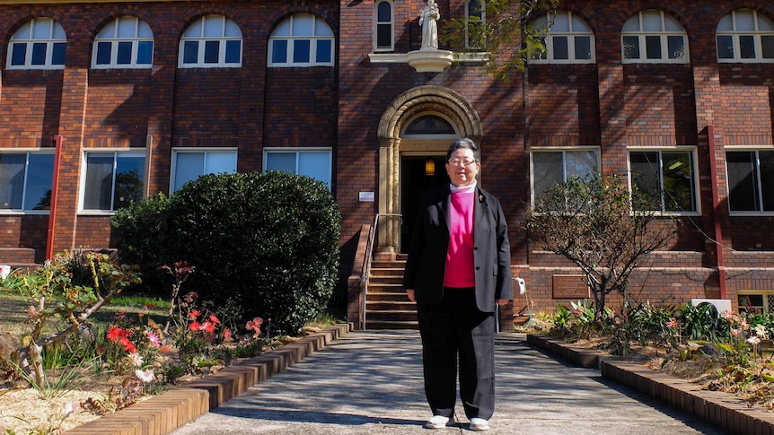 Josephite Sister Margaret Ng stands in front of brick religious building and garden.