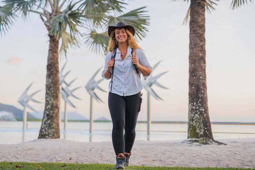 Photo of young woman with long hair, hat and backpack walking with beach, trees and Cairns Esplanade lagoon in the background