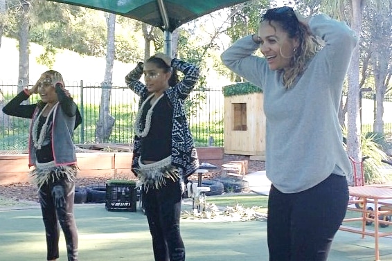 A young Indigenous woman with two Indigenous teen girls with their hands on their heads, smiling at a preschool.
