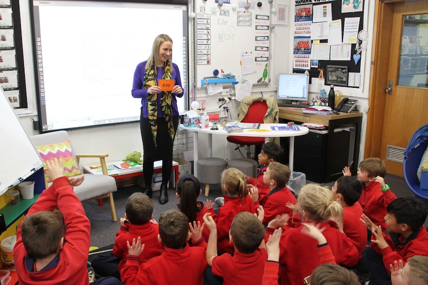 A teacher stands in front of a class of primary school kids holding a card showing the sound 'ew'.