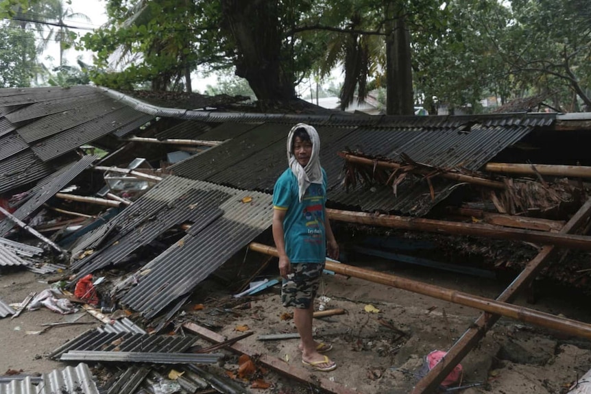 A man with a piece of fabric wrapped around his head stands among wreckage of corrugated iron with debris strewn everywhere.