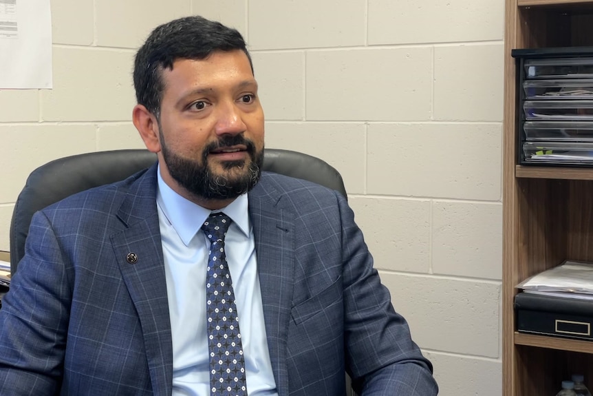 man with a beard sitting at a desk chair in a blue blazer