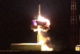 A missile launches at night, leaving a large exhaust blast of flames.