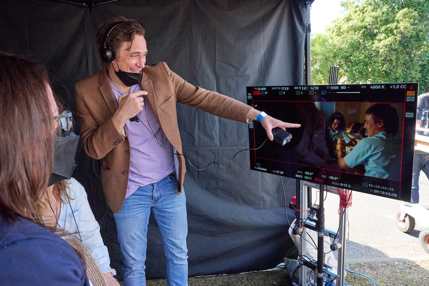 Trent smiles and points to a monitor behind the scenes of filming with plays a scene between the brothers