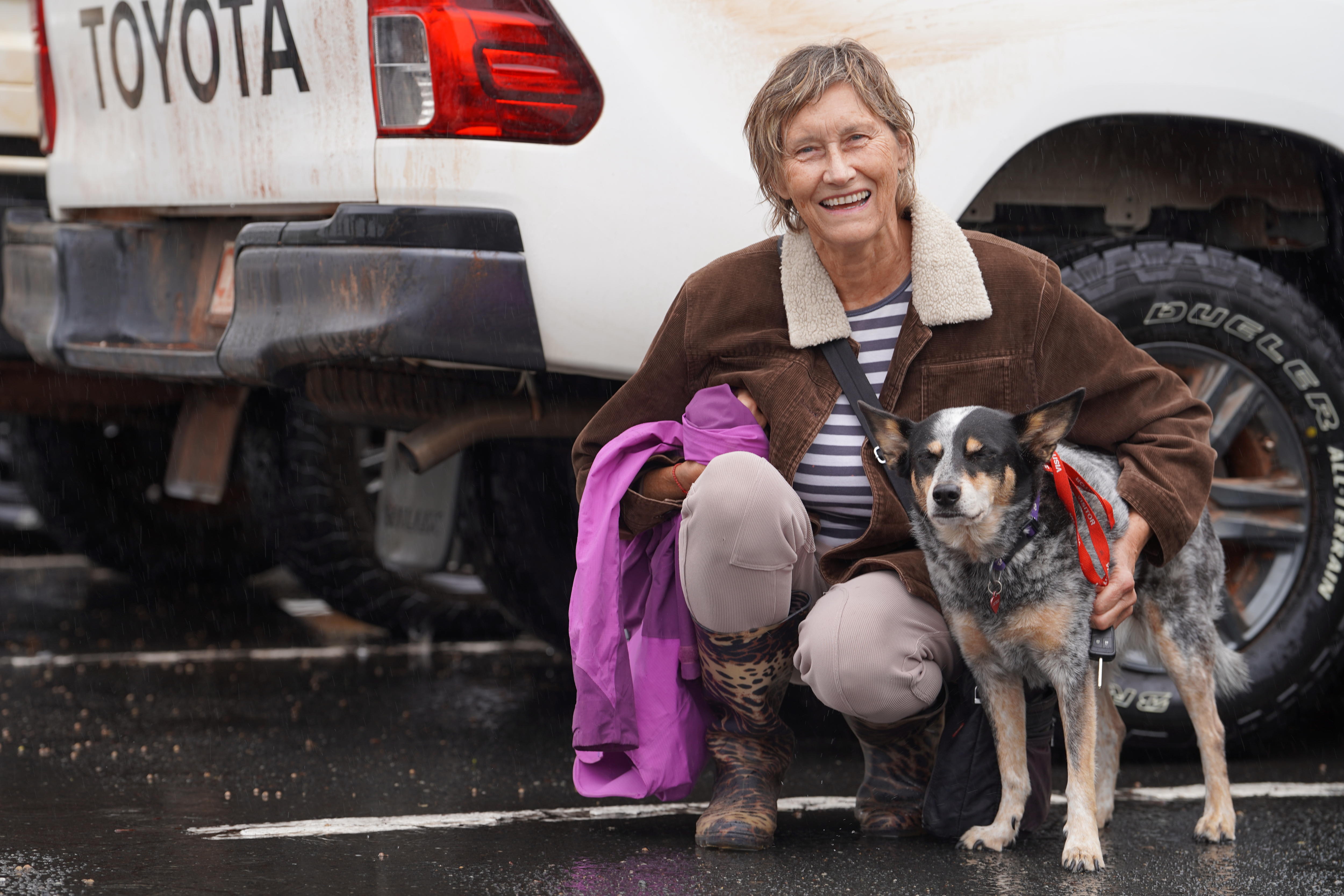 Woman squats near wet bitumen in front of 4WD with arm around border collie-looking dog 