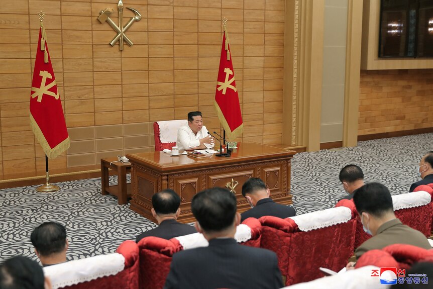 Kim Jong Un sits at a big, brown desk, which faces rows of men sitting in rows of red chairs 