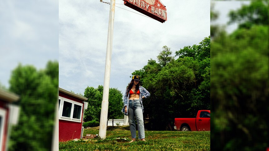 cover of Waxahatchee's 2024 album Tigers Blood, standing by a forestside truckstop in jeans, shirt and cap