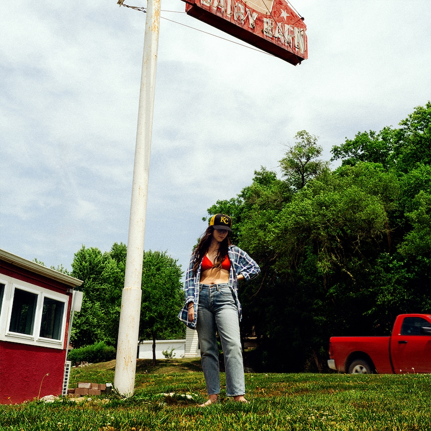 cover of Waxahatchee's 2024 album Tigers Blood, standing by a forestside truckstop in jeans, shirt and cap
