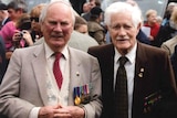 Two old men in suits, ties and a row of war medals smile for a photo with a crowd behind them.