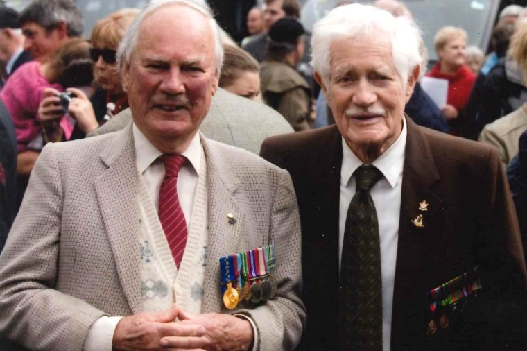 Two old men in suits, ties and a row of war medals smile for a photo with a crowd behind them.