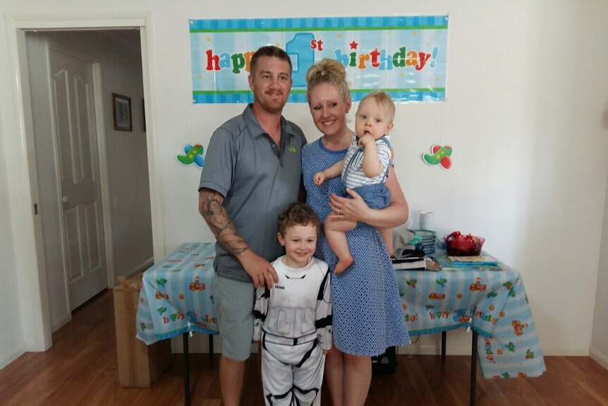 A husband and wife standing with a toddler and a baby in front of a happy birthday sign