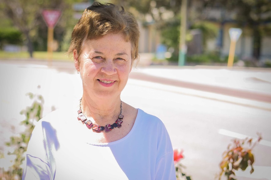 Older woman in white t-shirt stands outside in front of road