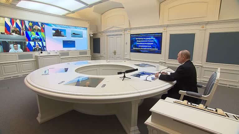 Vladimir Putin seated at white round table watches two screens with people and planes on it.