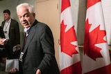 John McCallum looks back to the camera as he walks into a cabinet meeting as he is flanked by two Canadian flags on his right.