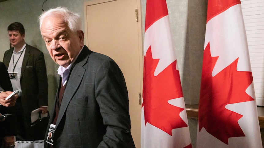 John McCallum looks back to the camera as he walks into a cabinet meeting as he is flanked by two Canadian flags on his right.