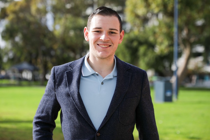 A young man with a blue shirt and blazer stands in a park smiling