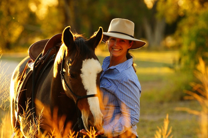 A woman in a hat smiling standing next to a horse holding its reins 