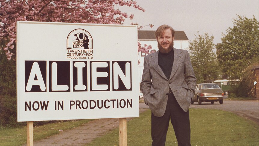 A man wearing suit stands outdoors next to a sign that reads 'Alien now in production' near a cherry blossom tree in a lot.
