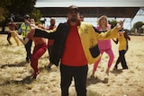 An Aboriginal man in hat and jacket stands arms out, a group of dancers - white and Aboriginal - behind him