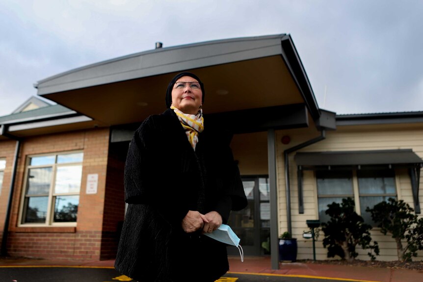 Woman in black coat and scarf wearing glasses stands outside aged care home beneath darkened skies