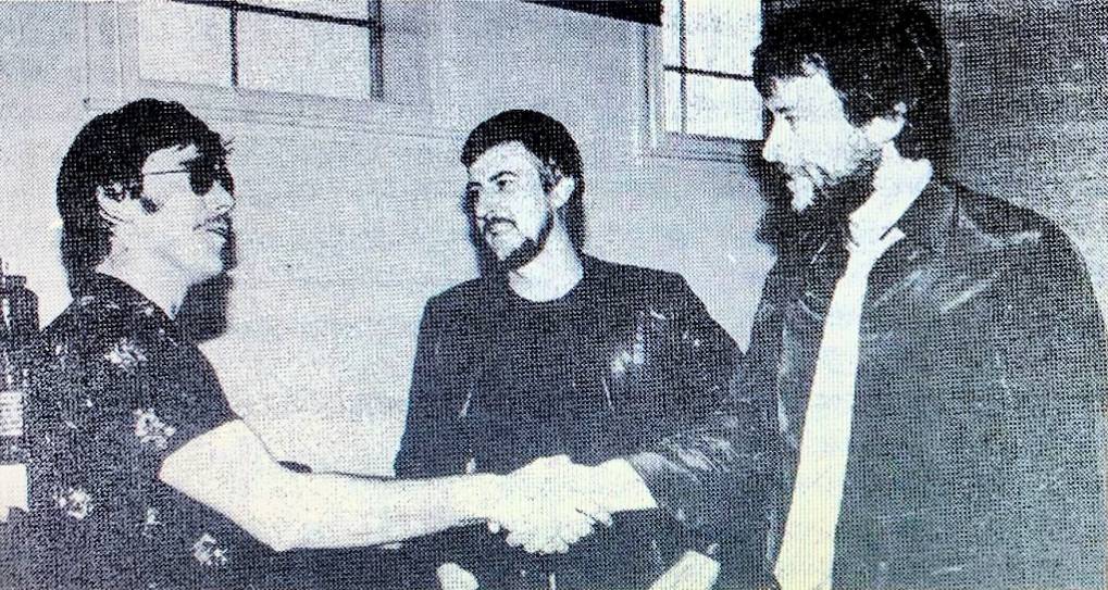 Una imagen en blanco y negro de dos hombres dándose la mano y otro hombre al fondo sobre un texto.