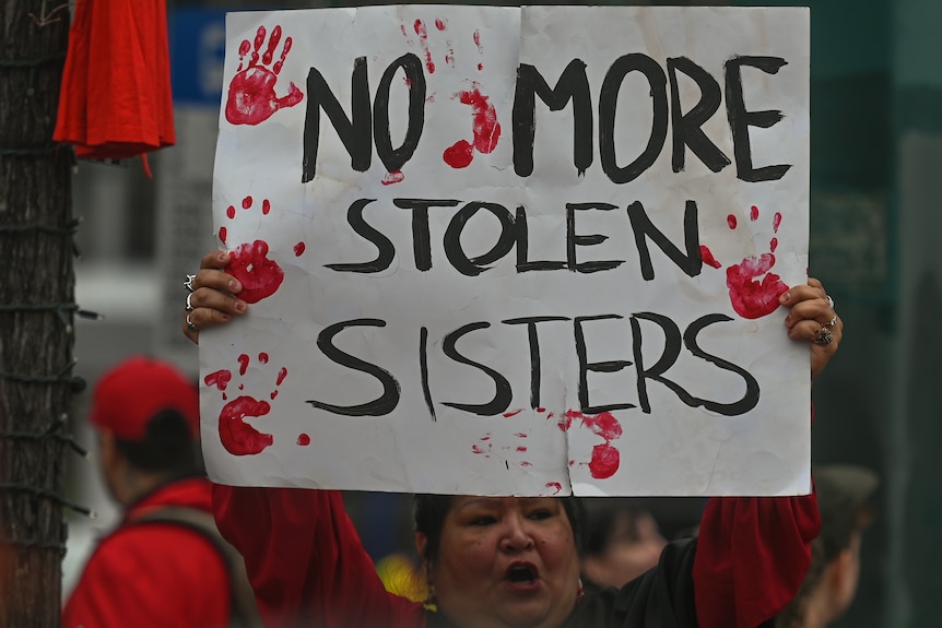 A woman holds a large placard above her head with the words, 'No More Stolen Sisters' with red hand prints around it.