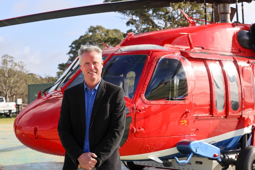 A man in a suit jacket stands in front of a red helicopter. 