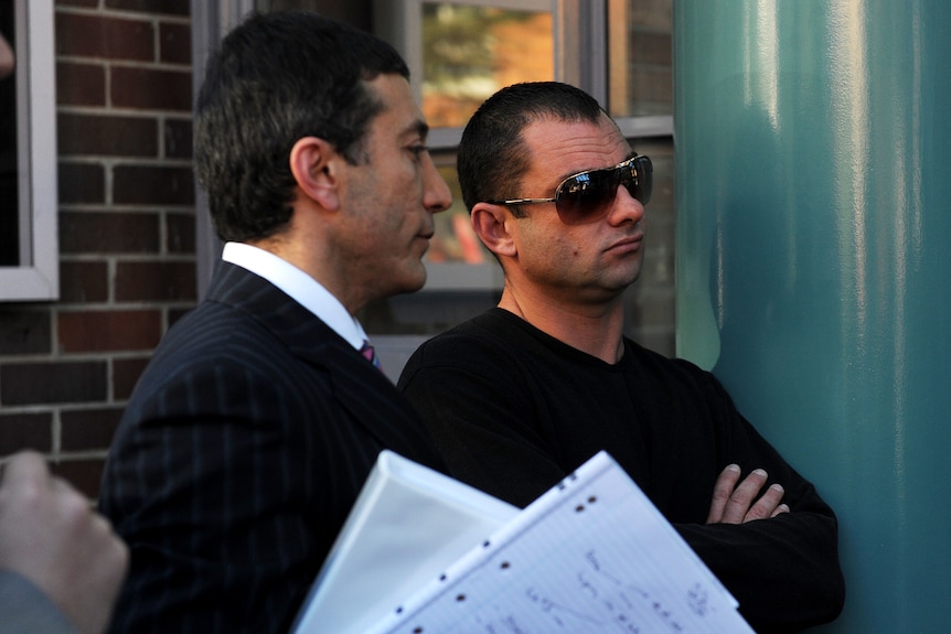 Man in black sunglasses stands with crossed arms next to man in suit with paper in the foreground.