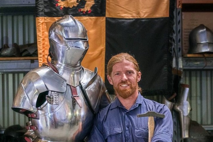 A man stands proudly besides medieval armour he has made.
