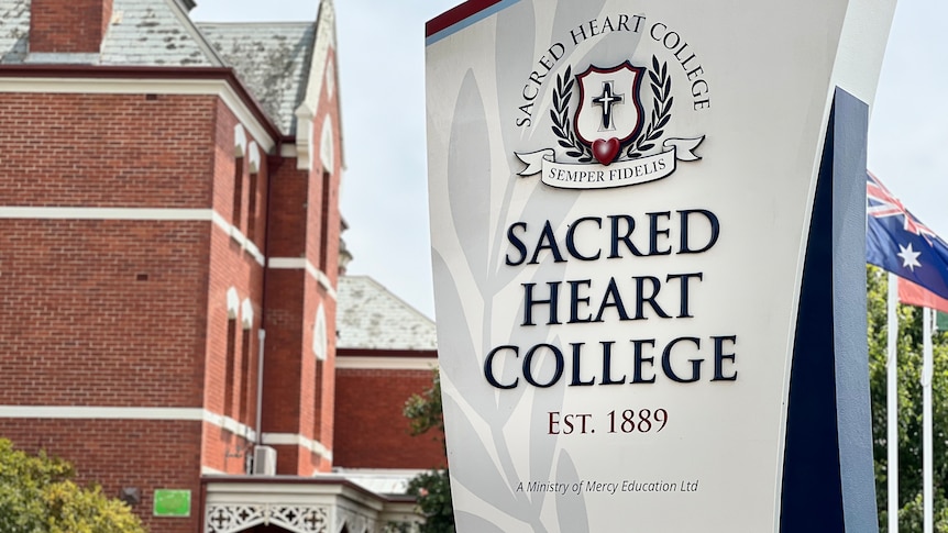 School sign that says Sacred Heart College with old red buildings behind.