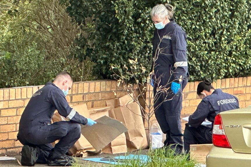 Two officers wearing blue forensics overalls kneel on a footpath and a third officer stands and watches as evidence is examined.