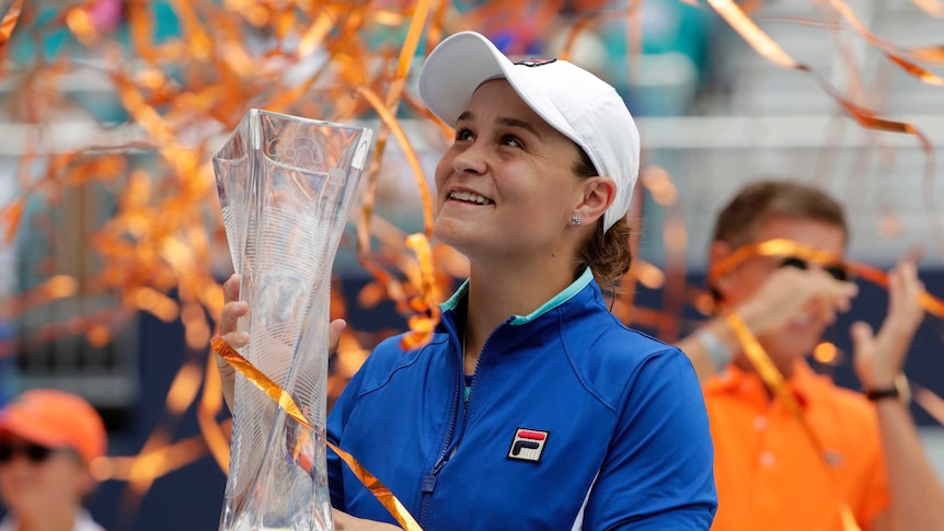 Ashleigh Barty looks up with a smile, holding a large, glass trophy in both hands while wearing a blue jacket and white cap