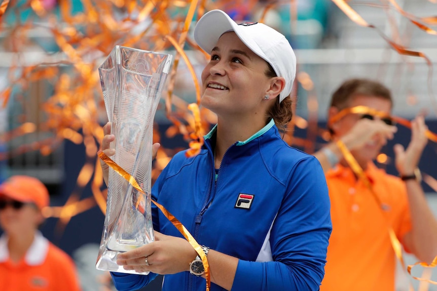 Ashleigh Barty looks up with a smile, holding a large, glass trophy in both hands while wearing a blue jacket and white cap