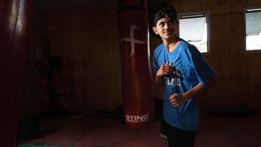 A young boy stands with his fists up in front of a heavy bag in a boxing gym.