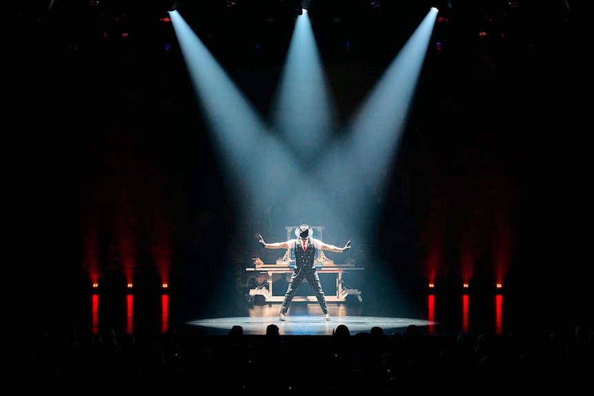 A man wearing hat on stage under lights looks at the floor