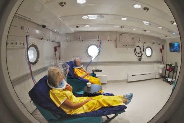 Two people in orange suits sit in chairs in a white hospital room 