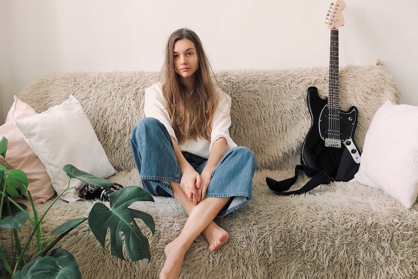 Young woman sitting on a shaggy couch surrounded by a guitar, pillows and indoor plant.