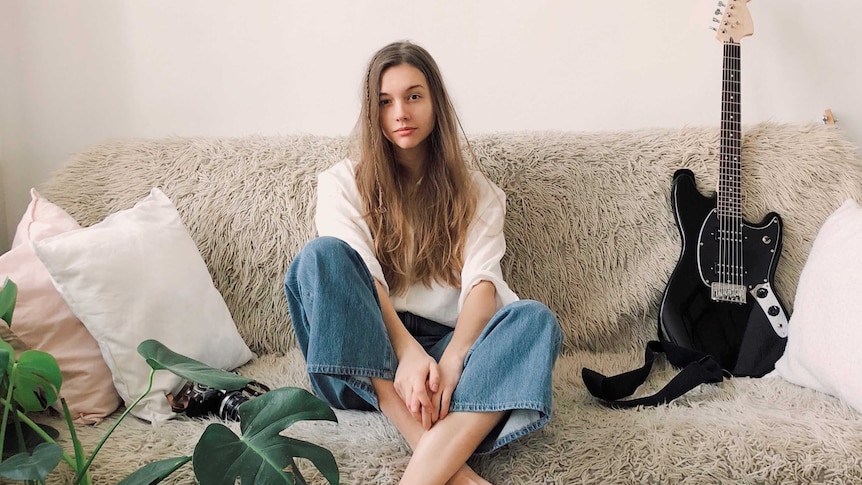 Young woman sitting on a shaggy couch surrounded by a guitar, pillows and indoor plant to depict finding your home style.