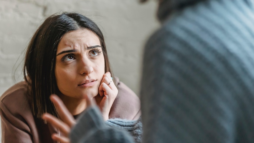 A woman wears a brown jumper and has her hand on her face as she looks up frowning while another person is seen talking to her. 