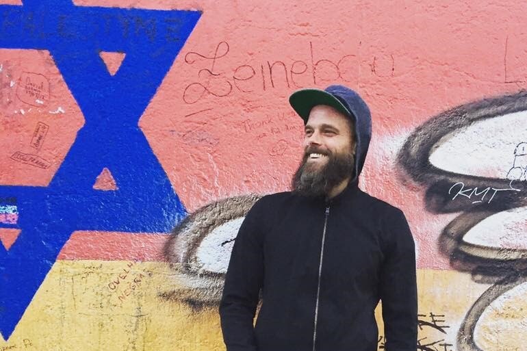 A bearded man in a hoodie smiles in front of street art.