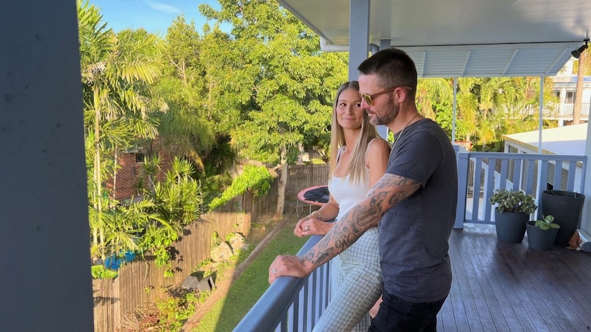 Taylor looks at Zane Warrell who is wearing sunglasses while looking out standing on their verandah.