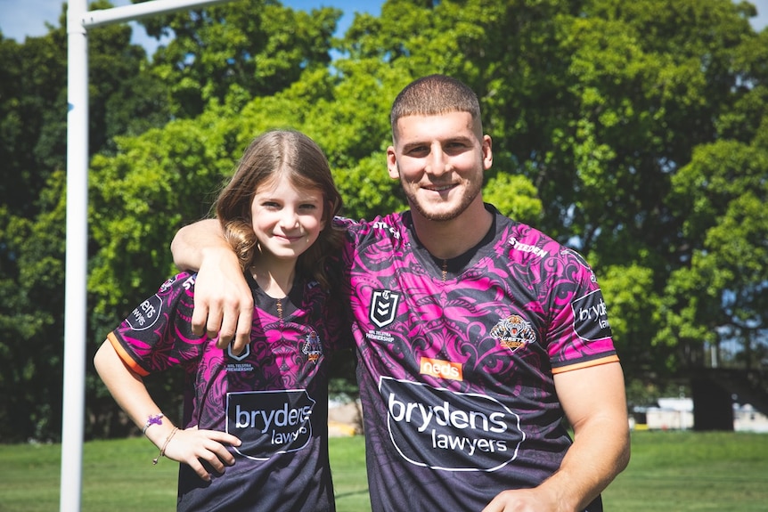 A young girl stands next to a rugby league player who has his arm around her. They're wearing pink jerseys and smiling.