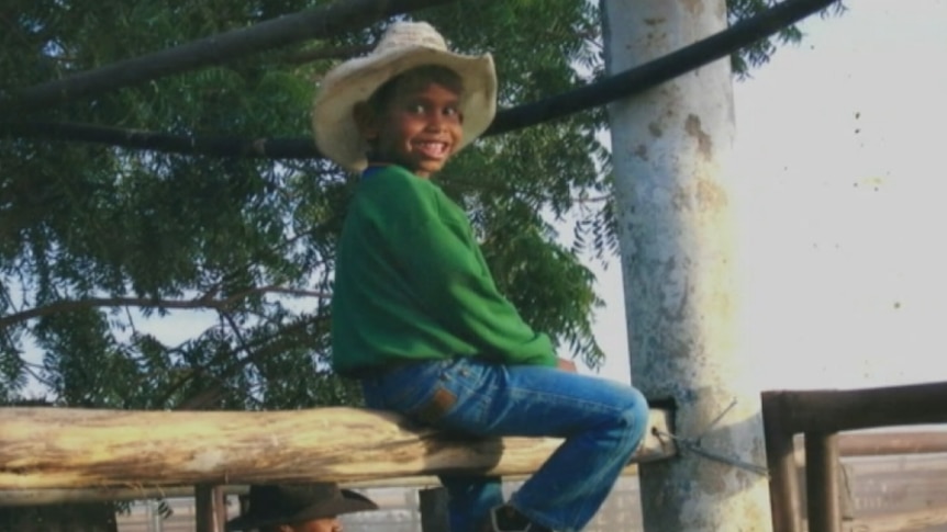 The child sits on a fence in blue jeans, a green jumper and a white cowboy hat