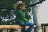 The child sits on a fence in blue jeans, a green jumper and a white cowboy hat