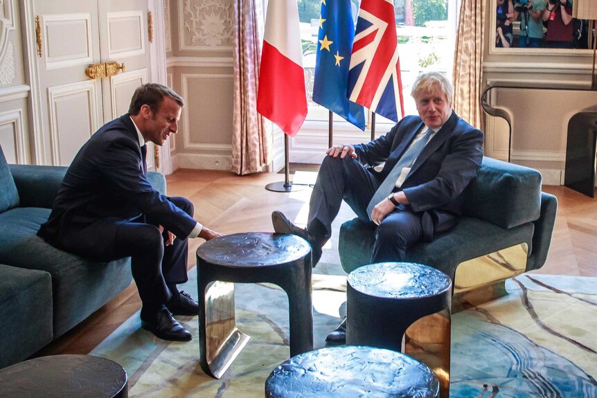 Boris Johnson puts his foot on a table as he sits across from Emmanuel Macron in a large room with a chandelier.