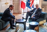 Boris Johnson puts his foot on a table as he sits across from Emmanuel Macron in a large room with a chandelier.