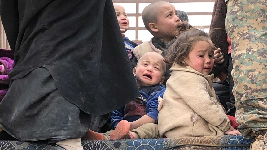 A group of children sit on the ground crying between the feet of two men, one in army fatigues.